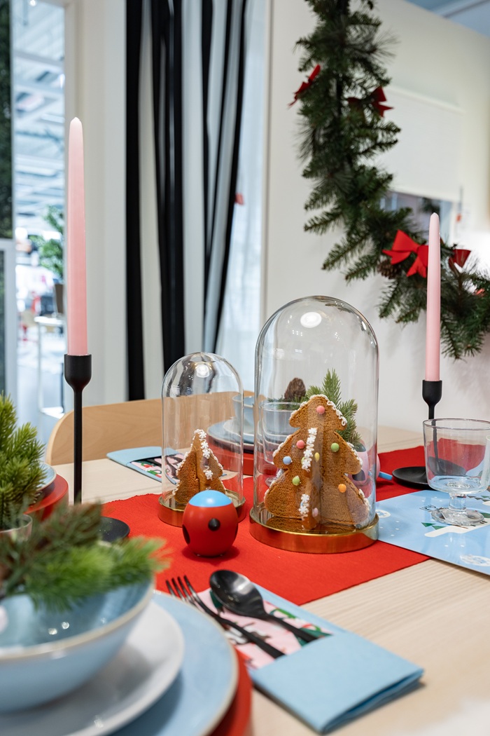 Punase linikuga laud, millel on sinised ja valged lauanõud. Klaaskupli all on piparkoogikuused, laual on mustas küünlajalas roosa küünal.        