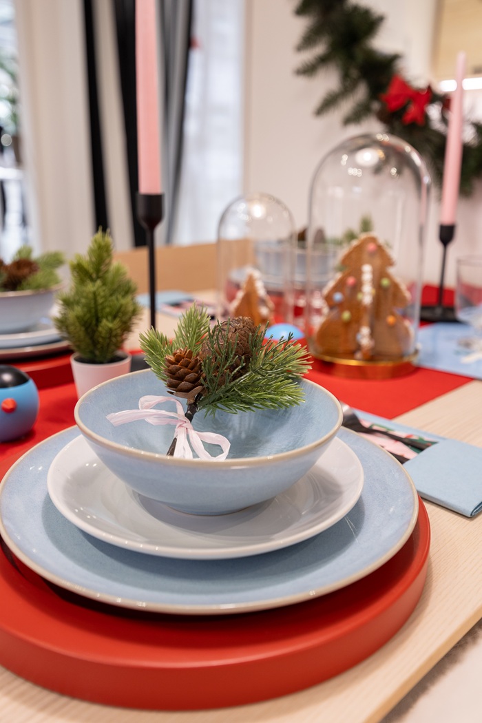 Lähivõte üksteise peale laotud valgetest ja heleisnistest taldrikutest, mis on asetatud punasele lauamatile. Ülemises kausis on väike roheline oks käbiga. Mustas küünlajalas on heleroosa küünal. 