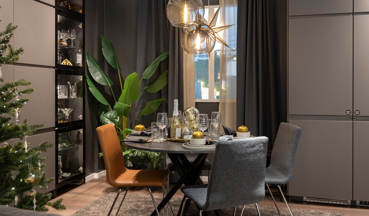 Kaasaegses toas on tume ümmargune laud, mida ümbritsevad hallid polsterdatud toolid ja üks pruuni nahaga tool. Toal on tumehallid seinad, siin on kõrge roheline taim ning hõbedaste ja kuldsete kaunistustega jõulupuu.