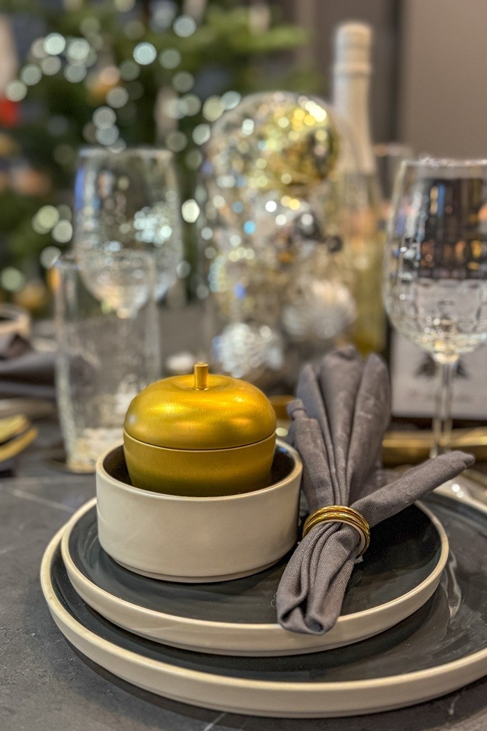 Lähivõte peolaual üksteise otsa laotud tumehallidest ja valgetest taldrikutest. Kõige peal on väike valge kauss, milles on särav kuldne õun. Nõude kõrval on hallist kangast salvrätik, mida hoiab koos kuldne rõngas.