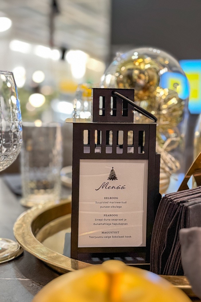 Tumepruun latern, milles on valge menüü, seisab kuldsel kandikul sädelevate kuldsete ja hõbedaste kaunistuste keskel. Hägustatud taustal on näha teised kaunistused.