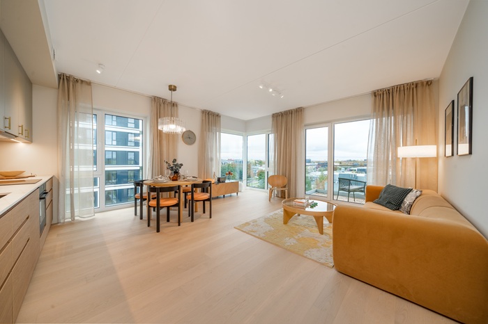 A spacious open living room with a dining area featuring dark wood table and chairs, a modern kitchen and beige curtains. The room has light-colored wooden floors.