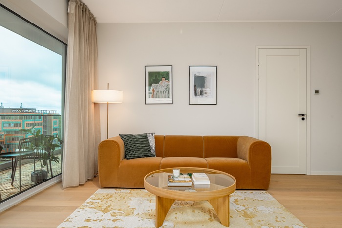 A living room area with a mustard-colored sofa, a round coffee table, and neutral-toned decor. A large window offers a view of an urban landscape.
