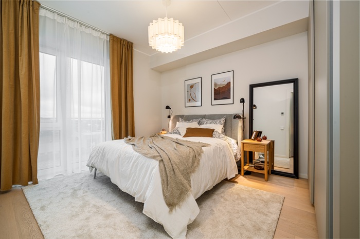 A spacious bedroom with warm gold curtains, light wood flooring and white and beige bedding.