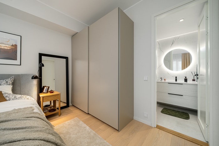 Gray wardrobes, neutral bedding, and a light wood nightstand in a spacious bedroom. A bright, modern bathroom with white surfaces and an illuminated mirror is visible through an open doorway.
