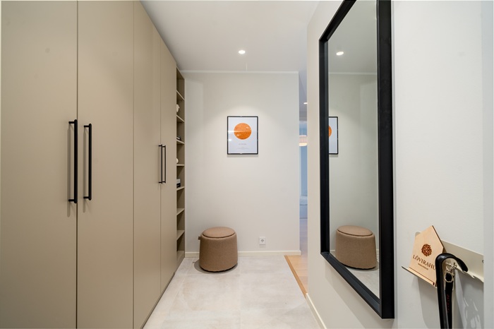 A hallway with beige wardrobes on the left and a large black-framed mirror on the right. On the far end, there´s a pouffe on the light colored floor.