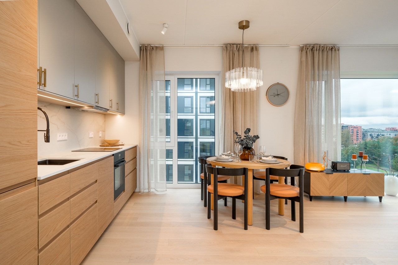 A modern kitchen with light wood cabinets, white countertops and a dining chairs with black frames and warm orange-brown seats. Large windows, beige curtains, and soft lighting enhance the airy, minimalist aesthetic.