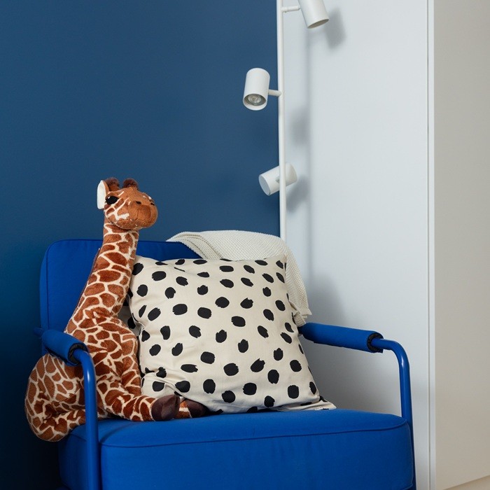 Plīša žirafe atrodas uz zila krēsla blakus baltam spilvenam ar melniem punktiem.