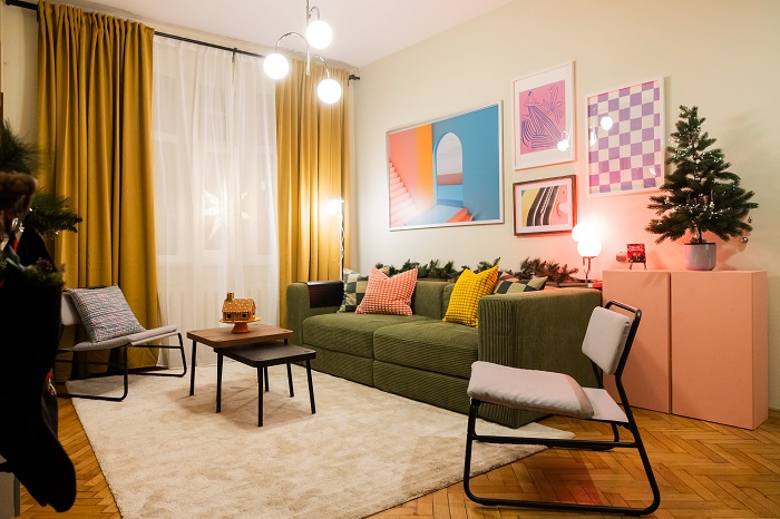 Krāsaina viesistaba ar dzelteniem aizkariem, atpūtas krēsliem, augiem un dekoratīviem attēliem pie sienas.