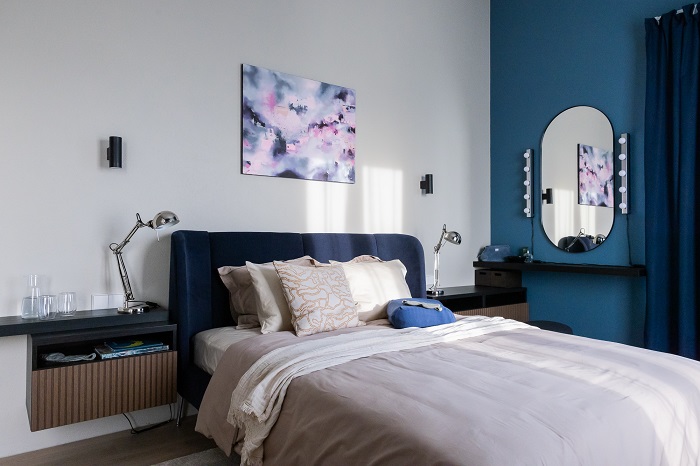 Mūsdienīga guļamistaba ar divguļamo gultu, zilām sienām, gaišu gultasveļu un vienkāršu apgaismojumu.