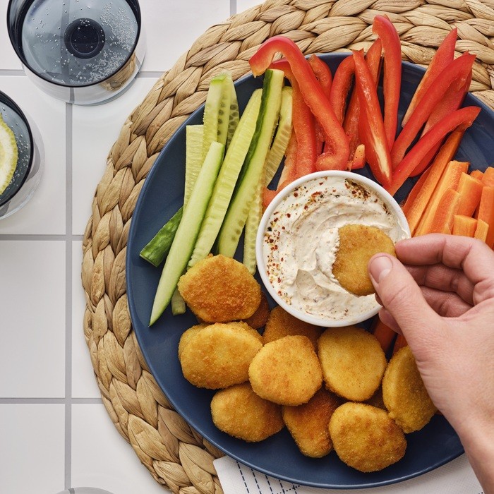 Ranka laiko augalinį kepsnelį ir merkia jį į indelį su padažu, o šalia, ant pinto stalo kilimėlio, guli šviežių daržovių lazdelės.