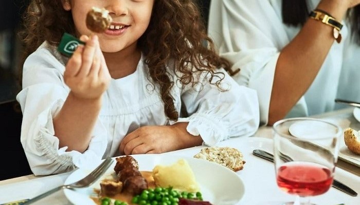 Vaikas sėdi prie stalo Švediškame restorane, laiko rankose augalinį kukulį ir šypsosi. Priešais, ant stalo, guli lėkštė su augaliniais kukuliais, bulvių koše, žirneliais ir bruknių uogiene.