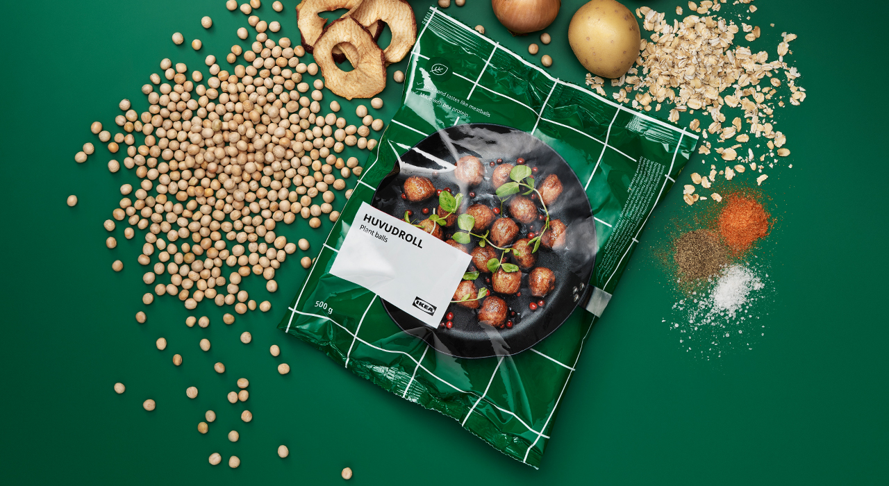 Iepakojums ar saldētām IKEA augu bumbiņām uz tumši zaļa fona, blakus izkaisīti produkti un garšvielas, kas ir to sastāvā.   