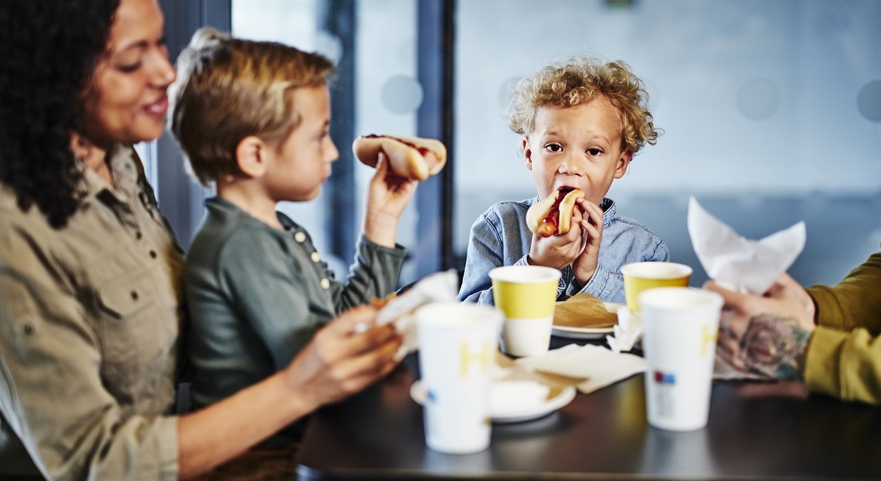 Divi bērni  un pieaugušais pie galda ēd hotdogus, uz galda atrodas kartona glāzes ar IKEA sveicienu Hej!.