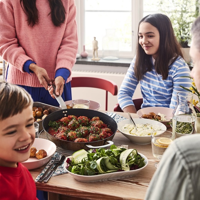 Ģimene, divi pieaugušie un divi bērni, pie galda ietur maltīti, sieviete servē pannā pagatavotas augu bumbiņas.