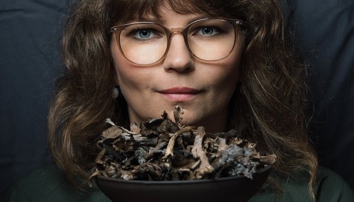 Lähivõte Maru Metspalust tumedal taustal, kus ta hoiab käes musta värvi kaussi, milles on erinevad taimsed koostisosad ja toiduained.