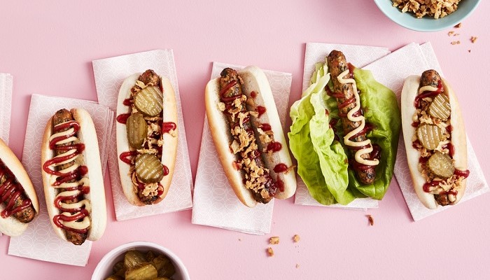 Erinevad hot dog'id koos lisanditega ülaltvaates, mis on asetatud roosale pinnale.