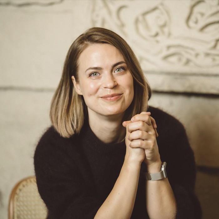 На фоне светлой кирпичной стены стоит улыбающаяся женщина в черном свитере, с короткими каштановыми волосами.