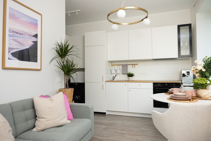 Gaiša viesistaba ar apvienotu virtuves zonu, baltiem skapīšiem, apaļu galdu, augiem un modernu griestu lampu.
