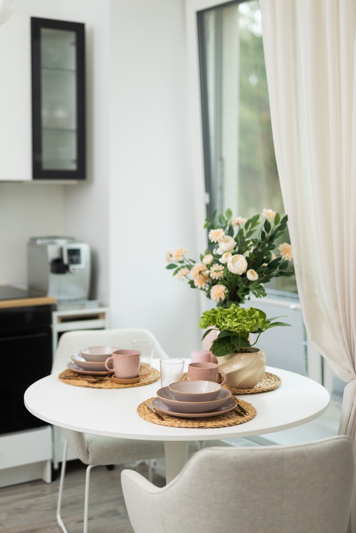 Apaļš galds pie loga ar klājumu, ziediem vāzē un aizkariem, radot mājīgu noskaņu.