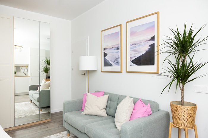 Gaiša atpūtas zona ar gaišu dīvānu, dekoratīviem spilveniem, ierāmētiem attēliem pie sienas un telpaugu stūrī.