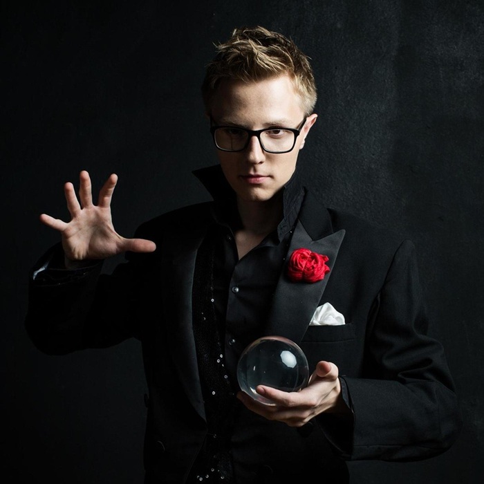 Мужчина-блондин в очках и чёрном костюме на тёмном фоне держит в одной руке стеклянный шар, а другую поднимает в жесте, будто показывает фокус.