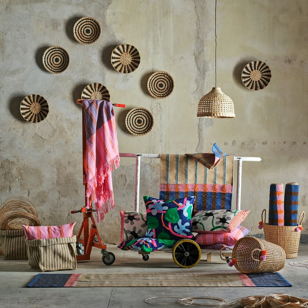 Green plates, bowls, and vases from the MÄVINN collection sit on a table in an industrial-style room. Two beige-green rugs lie beneath, and three plant pot hangers hang in the window.