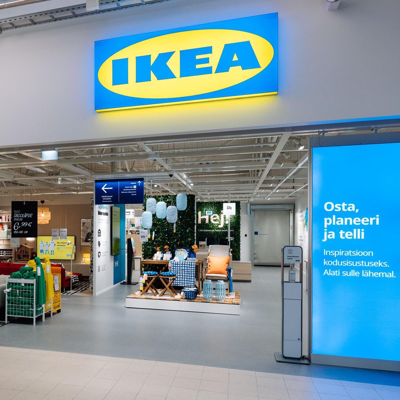Entrance to the new IKEA Pärnu small store in Estonia, with a large blue and yellow IKEA logo above. The entrance is open, showing a display of home furnishings and a brightly lit interior. On the right side, there is a large blue sign with text in Estonian. A hand sanitizer station stands next to the entrance.