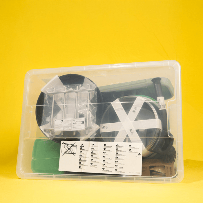 A transparent plastic storage box filled with various kitchen items, including pots, lids, utensils, and packaging materials. The background is bright yellow, and the box is sealed with tape and labels.