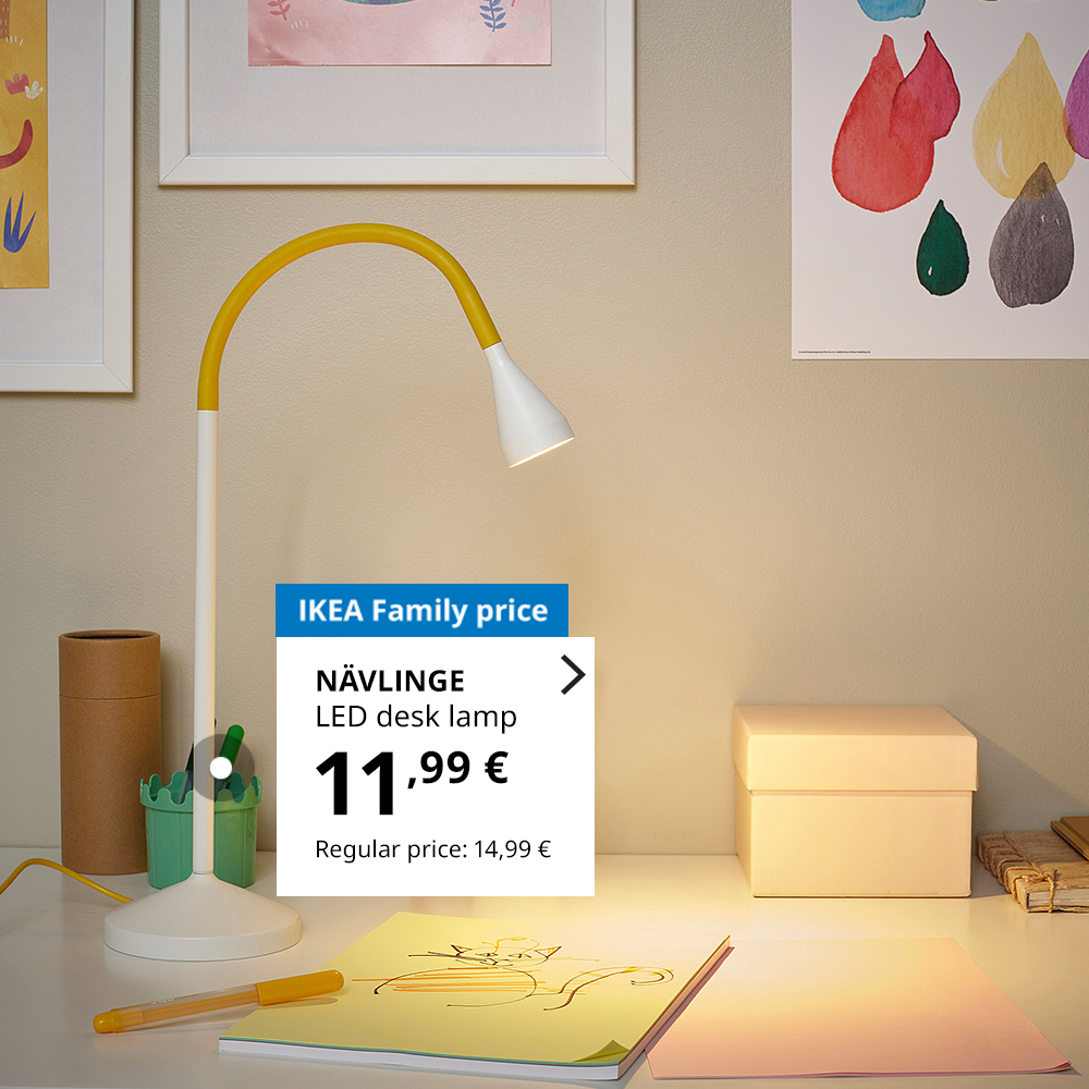 NÄVLINGE LED desk lamp with a flexible yellow neck placed on a desk in a child's room. On the desk are a drawing, a yellow pen, and a storage box. Colorful children's artwork is displayed on the wall. Price tag shows IKEA Family price — €11.99, regular price — €14.99.