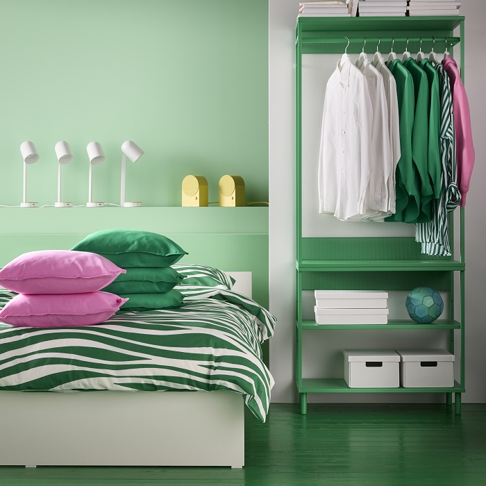A vibrant bedroom with a green and white striped bedding set, featuring pink and green cushions, an open shelving unit in green, some modern white table lamps and two yellow speakers.