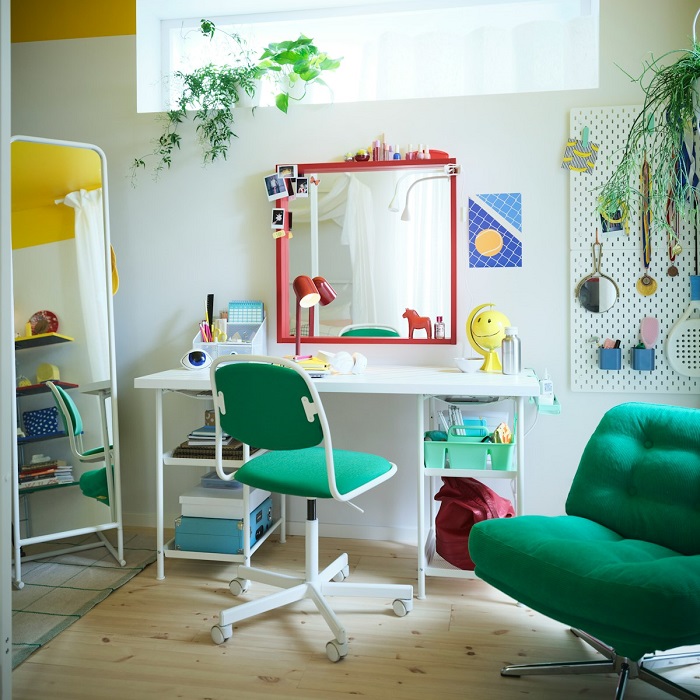 A bright children’s room with a white desk, green swivel chair, red-framed mirror, hanging plants, and a green armchair.