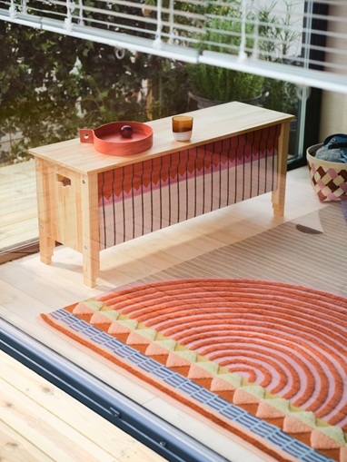 A handwoven OMMJÄNGE rug with a geometric pattern lies next to a wooden bench. The rug features a mix of short and long piles.