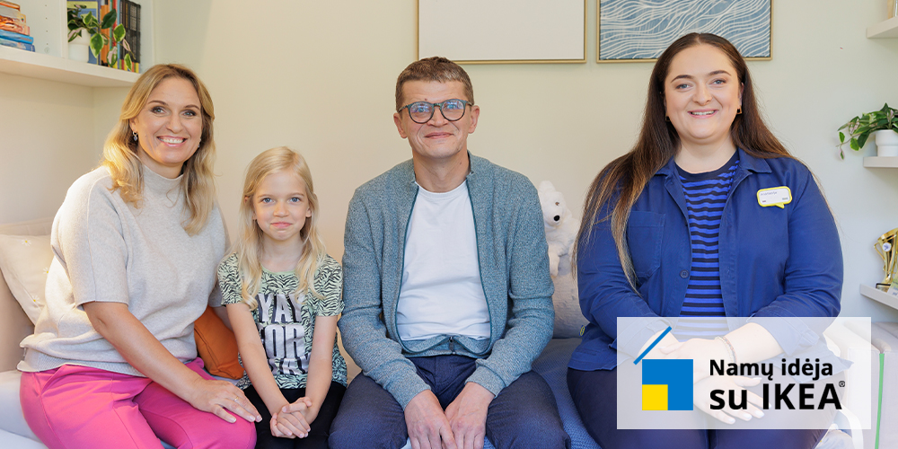 A family of three sits together on a bed in a cosy, well-lit room. The background shows a neatly arranged bookshelf and framed artwork on the wall.
