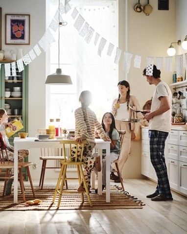 A bright, cosy kitchen with white cabinets, wooden chairs and a dining table, decorated with hanging garlands. Two adults and three kids are having a meal together.