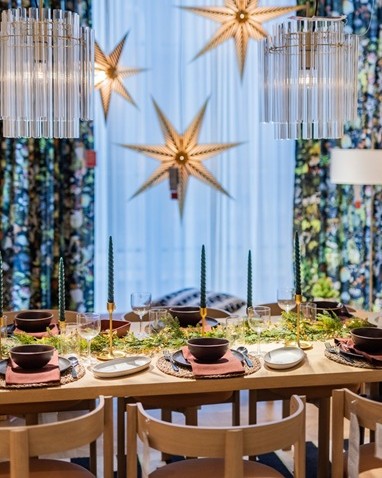 A festive dining table decorated with green candles, greenery and modern tableware, with large golden star ornaments hanging in front of patterned curtains.