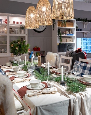 Vaade valge linaga peolauale. Sooja kumaga rippvalgustid tõstavad esile valgeid taldrikuid, rohelisi oksi ja punaseid salvrätte.