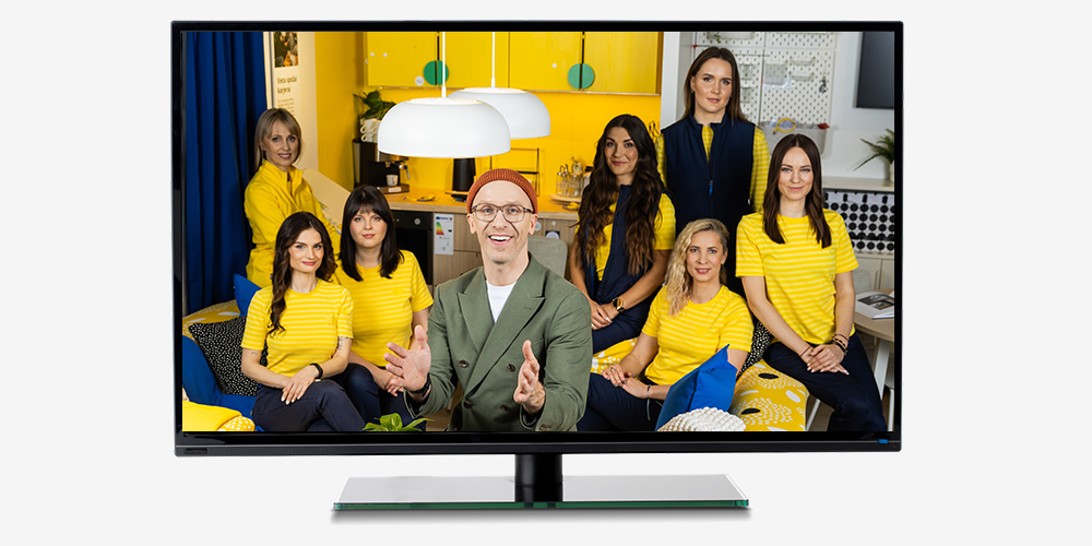 Ekrāns, kurā redzams, kā vīrietis zaļā uzvalkā sēž uz dzeltena dīvāna malas un smaida, apkārt viņam IKEA darbinieces formas apģērbā modernā, spilgti dzeltenā interjerā.