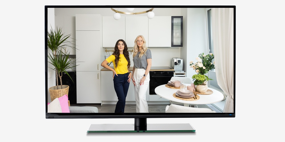 Televizora ekrānā redzamas divas sievietes, kas stāv gaišā virtuvē ar ēdamzonu; telpā ir balti skapīši, apaļš galds un telpaugi.