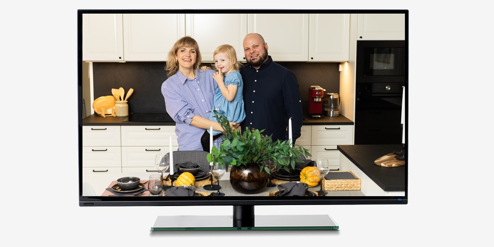 A screen showing family of three in a modern kitchen: a woman holding a young child with a man beside her. In the foreground is a dining table with black tableware, pumpkins, and green plants.