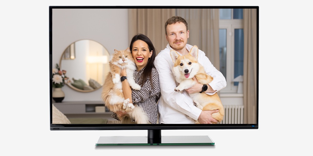 Ekrāns, kurā redzami sieviete un vīrietis gaišā guļamistabā, kuri tur rokās kaķi un suni, pozējot kamerai mājīgā un sirsnīgā atmosfērā.