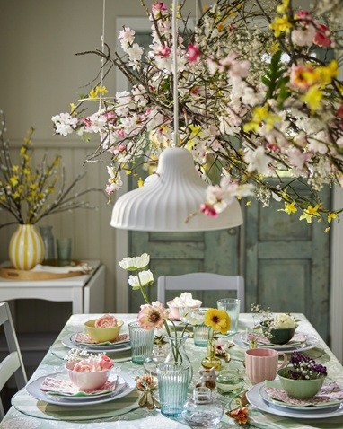 Pidulikult kaetud ja lilleoksakestega kaunistatud kevadine laud kollaste, tumeroheliste, roosade ja heleroheliste kaussidega, mis on asetatud väiksemate taldrikute peale, mis on omakorda suurematel taldrikutel.